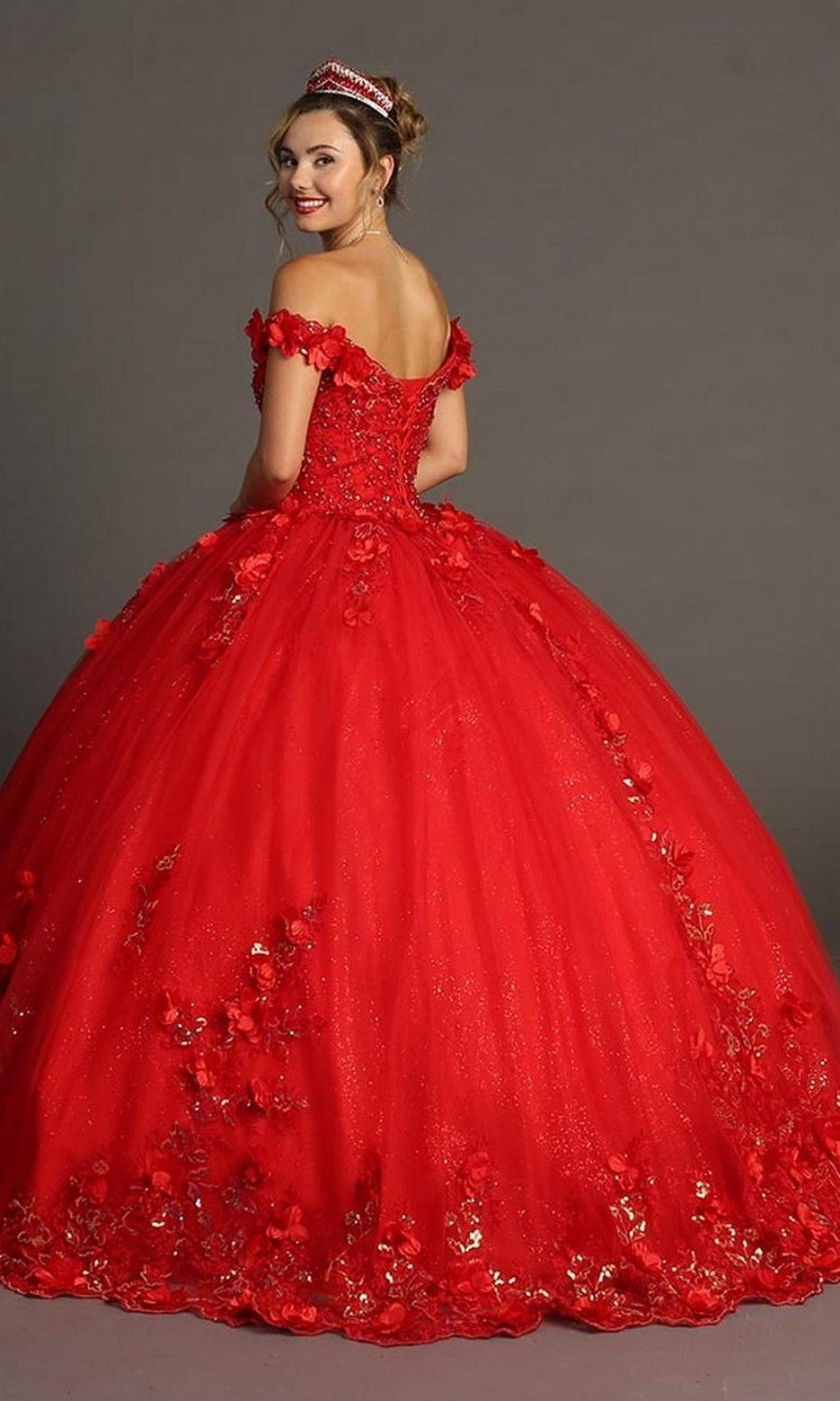 A young woman wearing the Quinceanera Dress CHF3085 By Chicas—a bright red, off-the-shoulder gown adorned with delicate floral appliques—stands facing away and smiles over her shoulder, her look completed with a sparkling tiara.